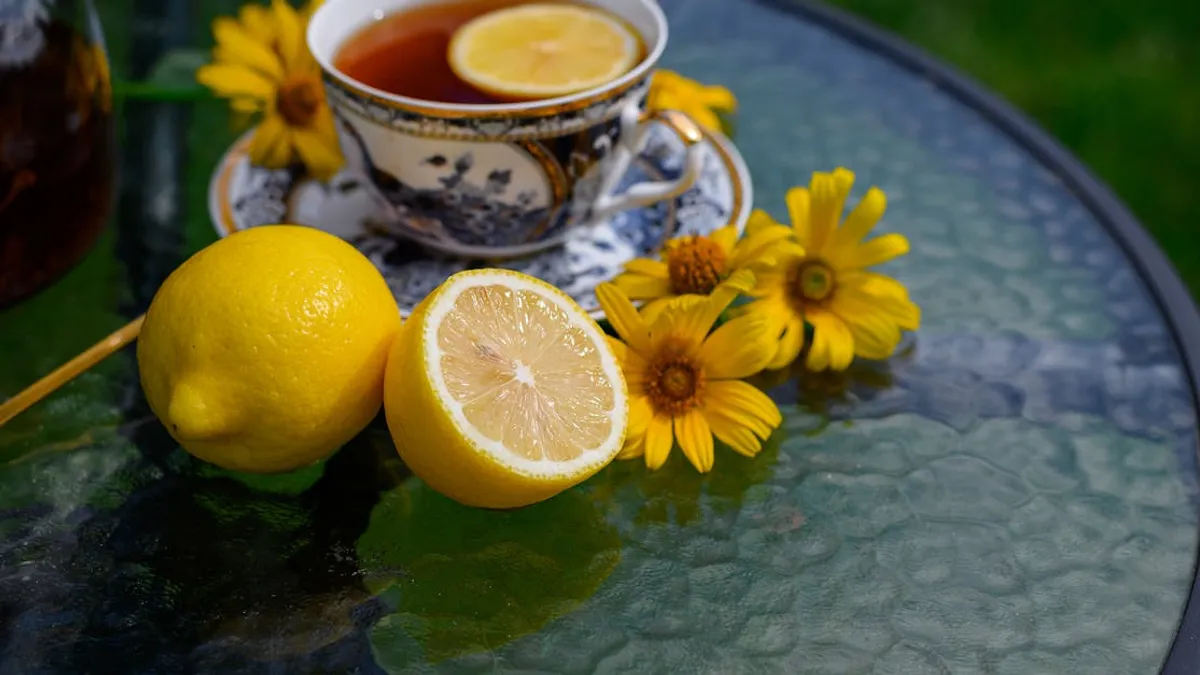 A refreshing cup of lemon tea on a glass table surrounded by yellow flowers and sliced lemon.
